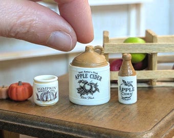 May include: Miniature ceramic apple cider and pumpkin themed decor. The set includes a small jug with a handle, a bottle, and a jar, all with labels. A tiny orange pumpkin is also present. The jug reads "Apple Cider" and "Farm Fresh".