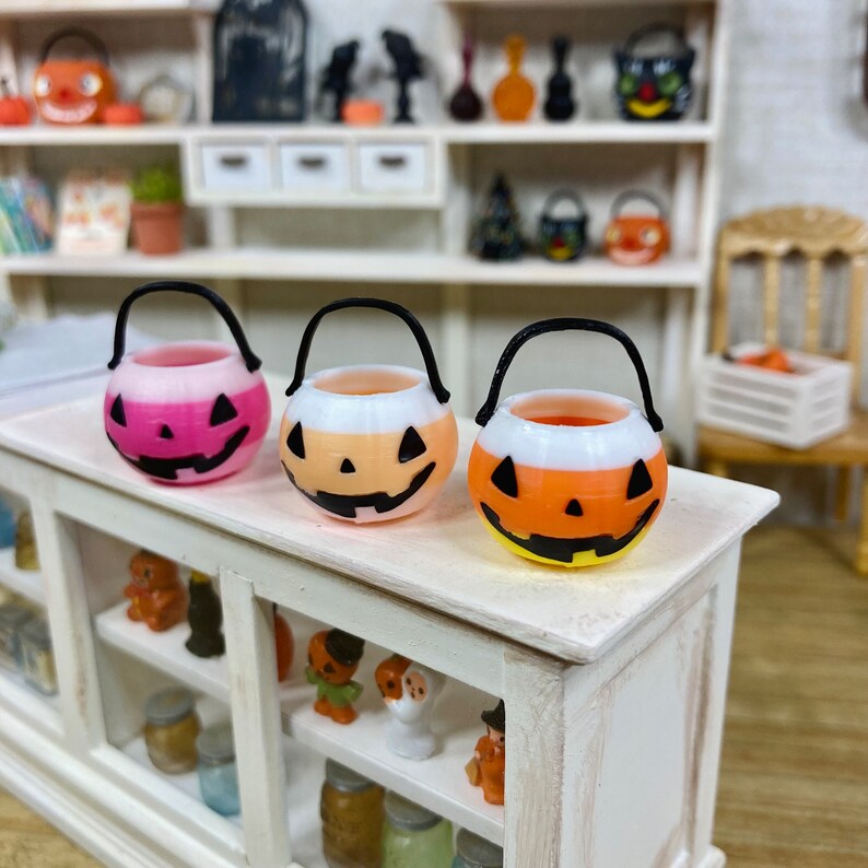 May include: Three miniature plastic jack-o'-lantern candy buckets with black handles. The buckets are decorated with candy corn stripes and have a white face with black eyes, nose, and mouth.