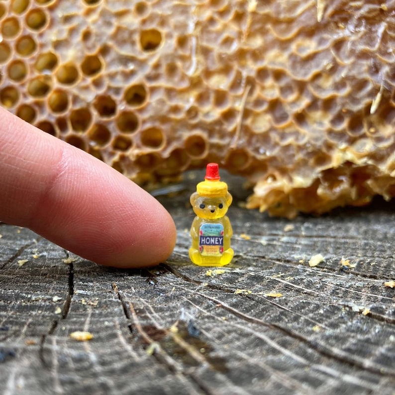 May include: A small yellow bear-shaped honey bottle with a red lid and the word "HONEY" printed on the front. The bottle is sitting on a wooden surface.