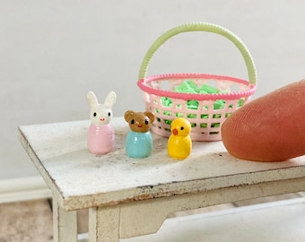 May include: Miniature Easter-themed figurines of a white bunny in a pink dress, a brown bear in a blue shirt, and a yellow duckling. A pink basket with a green handle and green contents sits behind the figurines on a white table.