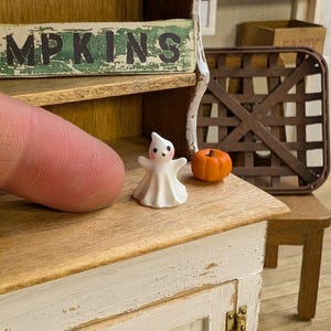 Pode incluir: Uma cena de Halloween em miniatura com uma pequena figura de fantasma branco e uma abóbora laranja. O fantasma tem os braços estendidos e um rosto sorridente. A palavra "MPKINS" é visível em uma placa de madeira acima.