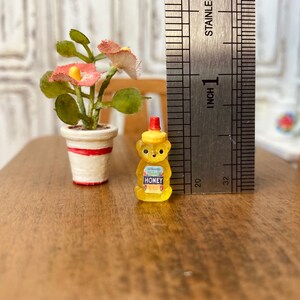 May include: A miniature honey bear bottle with a red lid and a white label that says "EVAPORATED HONEY" in blue lettering. The bottle is sitting on a wooden surface next to a miniature potted plant with red flowers.