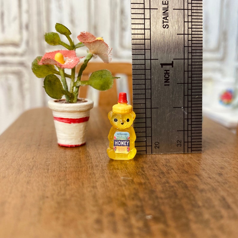 May include: A miniature honey bear bottle with a red lid and a white label that says "EVAPORATED HONEY" in blue lettering. The bottle is sitting on a wooden surface next to a miniature potted plant with red flowers.