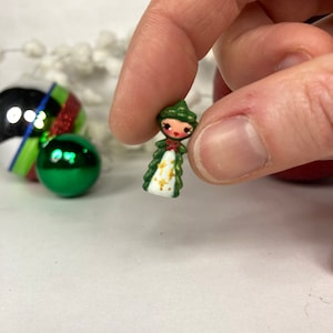 May include: A small Christmas tree-shaped ornament, held between fingers. The doll has a green hat and dress with gold detailing. Other Christmas ornaments are in the background, including a green and red striped ball.