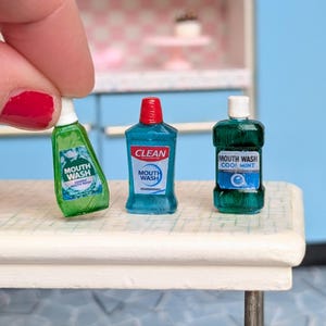 May include: Miniature mouthwash bottles in green and blue with white labels. One bottle is held by a hand with red nail polish. The bottles have labels that read "Mouth Wash" and "Cool Mint". The bottles are on a white surface.
