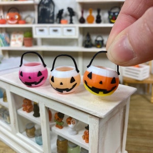 May include: Three miniature Halloween trick-or-treat buckets shaped like jack-o'-lanterns. The buckets are pink, orange, and yellow with black eyes, nose, and mouth details. The buckets have black handles.