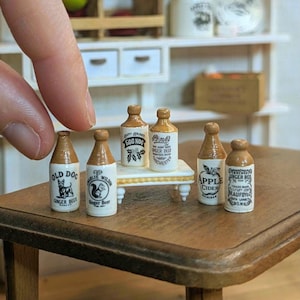 Pode incluir: Miniaturas de garrafas de cerveja de gengibre com tampas castanhas e rótulos brancos. Os rótulos apresentam texto e ilustrações, incluindo "Old Dog Ginger Beer" e "Apple Cider". As garrafas estão dispostas numa pequena mesa de madeira.