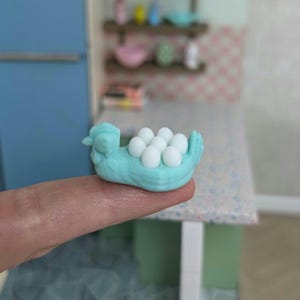 Pode incluir: Uma figurinha de frango de cerâmica azul claro segurando um ninho de ovos brancos. O frango está sentado em um dedo.