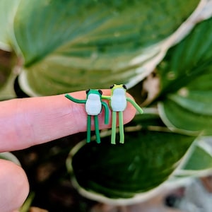 Puede incluir: Dos pequeñas figuras de rana hechas a mano. Una rana es verde con vientre blanco y la otra verde claro con vientre blanco. Ambas tienen ojos negros y posan con los brazos extendidos. El fondo son hojas verdes.