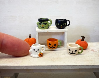 May include: Five miniature Halloween-themed mugs on a white wooden table. The mugs are decorated with a ghost, a pumpkin, a black cat, and a Frankenstein. Two small orange pumpkins are also on the table.