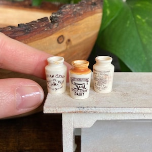 Puede incluir: Tres mini botellas de leche de cerámica con etiquetas. Las botellas son blancas con letras e imágenes marrones. Las etiquetas dicen "Avonah Cream Dairy", "Daisy Acres Farm Thick-Cream Dairy" y "Anne & Shirleys Primrose Dairy".