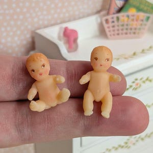 May include: Two miniature baby dolls with peach-coloured skin and rosy cheeks. The dolls have small, detailed features, including closed eyes and tiny limbs. One doll is seated, and the other is standing. The dolls are likely for a dollhouse or miniature collection.