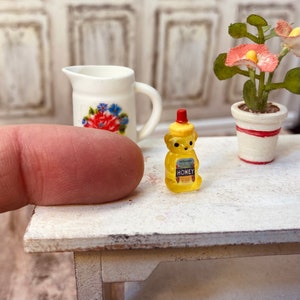 May include: A miniature yellow bear-shaped honey bottle with a red lid and a white label that says "HONEY". The bottle is sitting on a white wooden table.