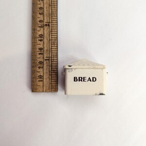 May include: A miniature off-white metal bread box with a lid and the word BREAD printed in black on the front. A wooden ruler is positioned next to the item, showing it measures approximately 1.5 inches in height and 2 inches in width.