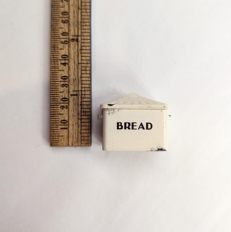 May include: A miniature off-white metal bread box with a lid and the word BREAD printed in black on the front. A wooden ruler is positioned next to the item, showing it measures approximately 1.5 inches in height and 2 inches in width.