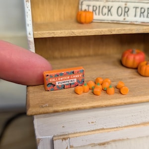 May include: Miniature Halloween-themed display featuring an orange box of "Halloween Candy Pumpkins" and several small orange pumpkin candies. A finger holds the candy box. Other miniature pumpkins are on a shelf.