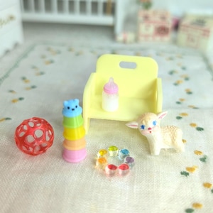 May include: A miniature scene featuring a yellow chair with a baby bottle, a colorful stacking toy, a red ball, a rainbow ring, and a small lamb figurine. The toys are arranged on a patterned surface, creating a playful and whimsical aesthetic.
