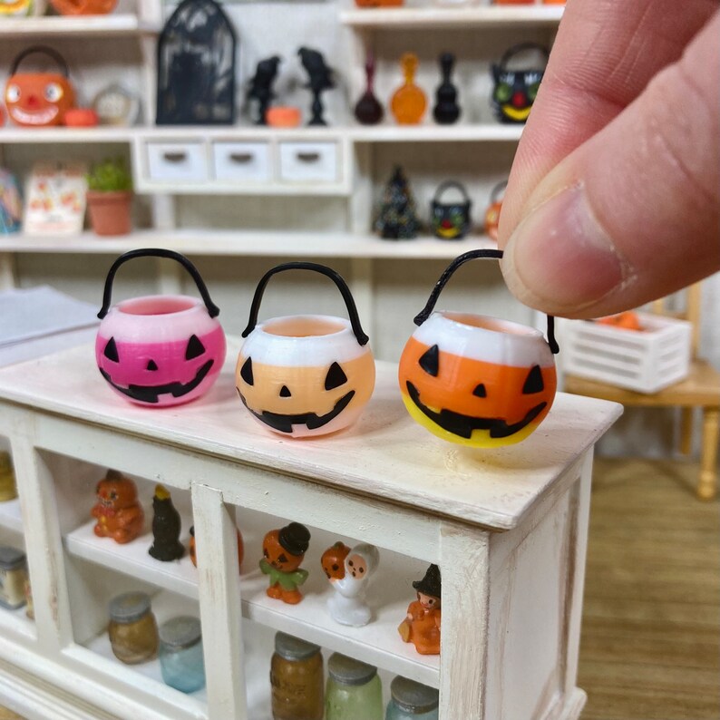 May include: Three miniature Halloween trick-or-treat buckets. One is pink and white, one is orange and white, and one is yellow and white. Each bucket has a black handle and a jack-o'-lantern face.