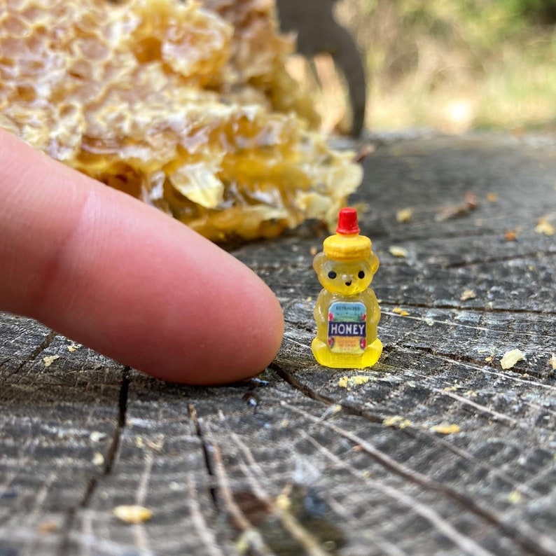 May include: A small yellow bear-shaped honey bottle with a red lid. The bottle is sitting on a wooden surface with a honeycomb in the background.