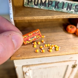 May include: Miniature Halloween candy box with the words "HALLOWEEN CANDY CORN" and scattered candy corn pieces. The box is orange and yellow. The candy corn pieces are yellow, orange, and white.