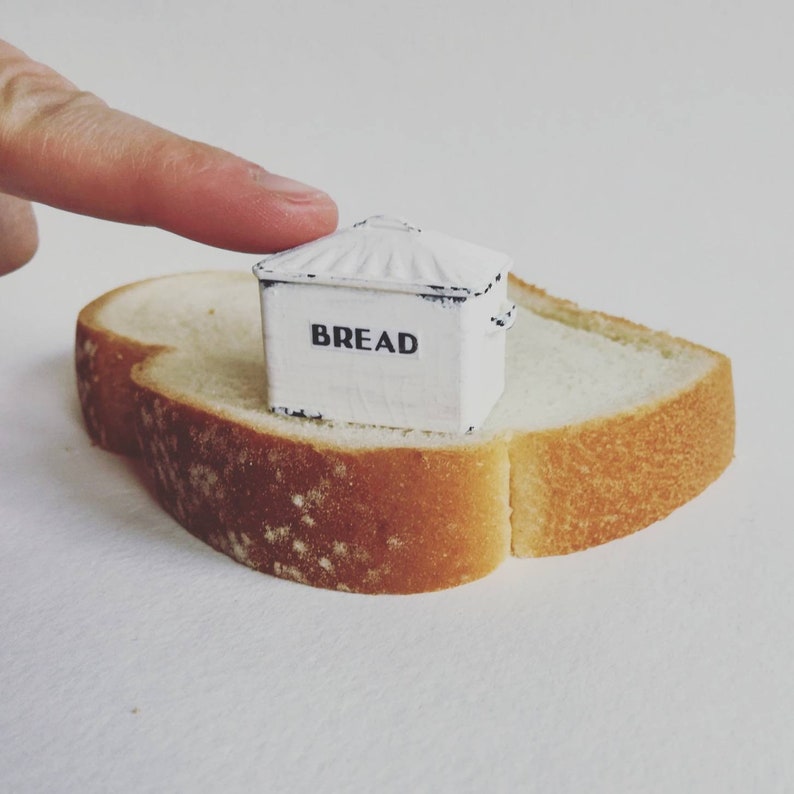 May include: A miniature white metal bread box with a distressed finish and a slanted lid, featuring the word BREAD printed in black capital letters on the front, resting on a single slice of white bread with a golden brown crust edge.