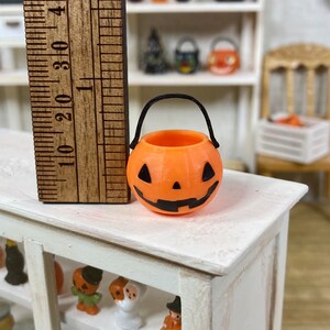 May include: A small orange pumpkin-shaped Halloween trick-or-treat bucket with a black handle. The bucket has a carved jack-o'-lantern face.