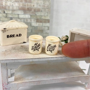 May include: Two miniature white ceramic canisters with black lettering. The canisters are labeled "Sugar" and "Flour".