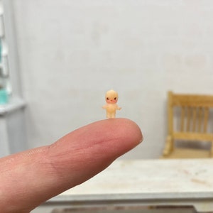 Peut inclure: Une petite poupée jaune pâle avec des lèvres et des yeux rouges peints se tient sur un doigt. La poupée porte une simple robe blanche et a une petite tête ronde.