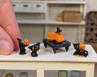 Może przedstawiać: Miniaturowe figurki na Halloween. Kolekcja obejmuje czarnego kota, figurkę z dyniową głową i pomarańczowego ducha w kapeluszu czarownicy na czarnym stole. Postać w czarnym płaszczu trzyma dynię. Scena rozgrywa się na białej półce.