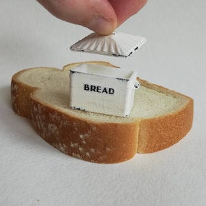 May include: A miniature white rectangular bread box with black distressed edges and the word BREAD printed in black on the front. The box has a removable lid with a ridged texture, and it is positioned on top of a single slice of white bread.