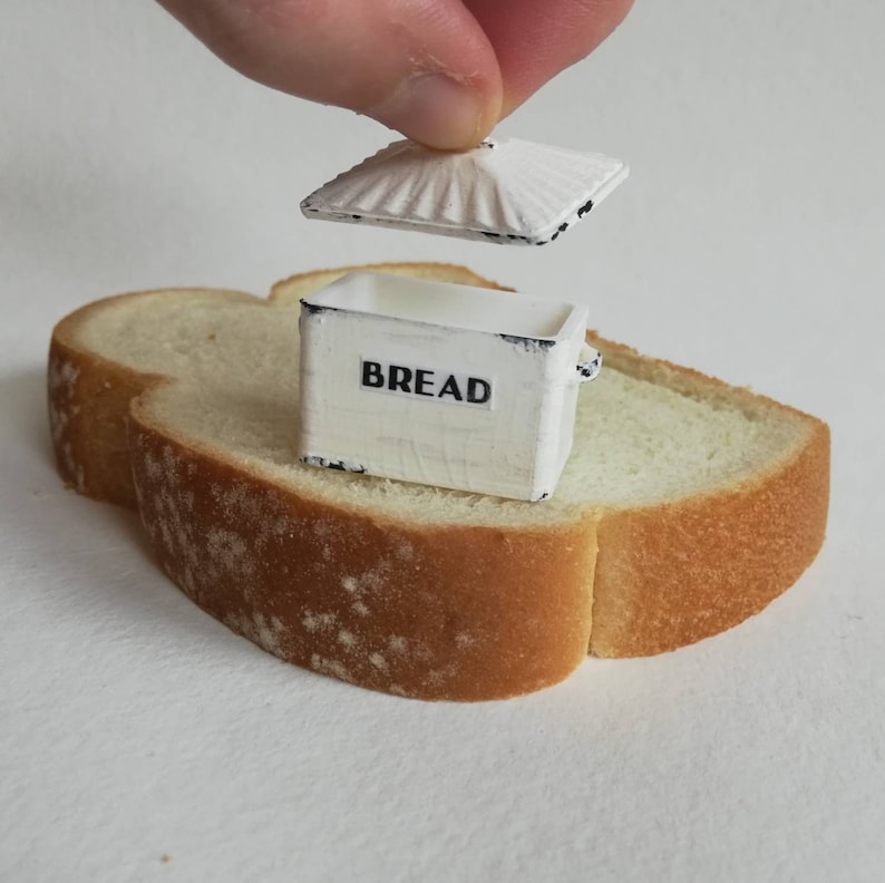 May include: A miniature white rectangular bread box with black distressed edges and the word BREAD printed in black on the front. The box has a removable lid with a ridged texture, and it is positioned on top of a single slice of white bread.