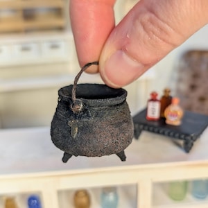 Puede incluir: Caldero negro en miniatura con asa, apoyado sobre tres patas pequeñas. El caldero tiene un aspecto texturizado y envejecido. Una pequeña mesa con botellas está en el fondo. El caldero está sostenido por los dedos de una persona.