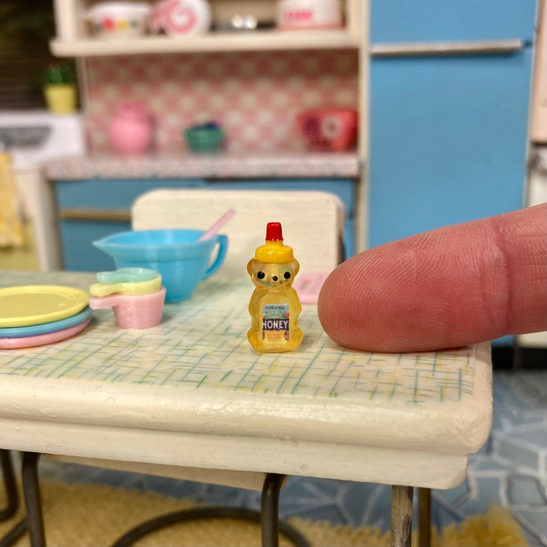 May include: A miniature yellow honey bear bottle with a red lid and the word "Honey" printed on the label. The bottle is sitting on a white table with a checkered pattern.