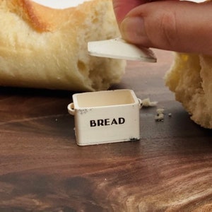 May include: A miniature off-white metal bread box with a removable lid, featuring the word BREAD printed in black sans-serif font on the front. The small container has two side handles and shows subtle distressed detailing along the edges.