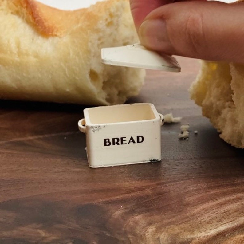May include: A miniature off-white metal bread box with a removable lid, featuring the word BREAD printed in black sans-serif font on the front. The small container has two side handles and shows subtle distressed detailing along the edges.