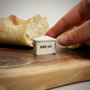 May include: White ceramic bread box with the word "BREAD" printed on the front. The box is sitting on a wooden surface next to a loaf of bread.
