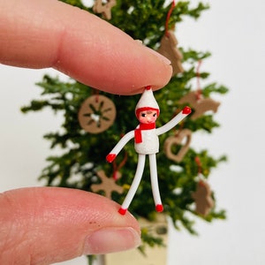 May include: A miniature Christmas elf figurine with a white body, red accents, and a white hat. The elf is held between fingers, with a small Christmas tree and gingerbread ornaments in the background. The elf is approximately 1 inch tall.