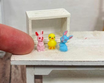 May include: Three miniature figurines: a pink bunny, a yellow chick, and a blue bunny. The figurines are arranged in a row on a white wooden table.