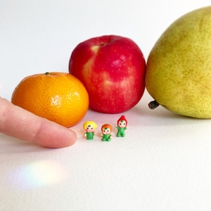 May include: Three miniature figurines standing in a row, each wearing a green outfit. One has a yellow hat, one has an orange hat, and one has a red pointed hat. They are positioned next to a bright orange tangerine, a red apple, and a green pear.