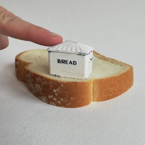 May include: A miniature white metal bread box with a distressed finish and a ridged lid, featuring the word BREAD printed in black on the front. The small container is resting on a single slice of white bread with a golden brown crust.