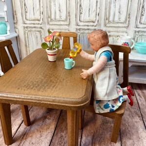 May include: A doll wearing a white dress with blue floral print is pouring yellow liquid from a bottle into a blue cup. The doll is sitting at a wooden table with a potted plant with pink flowers.