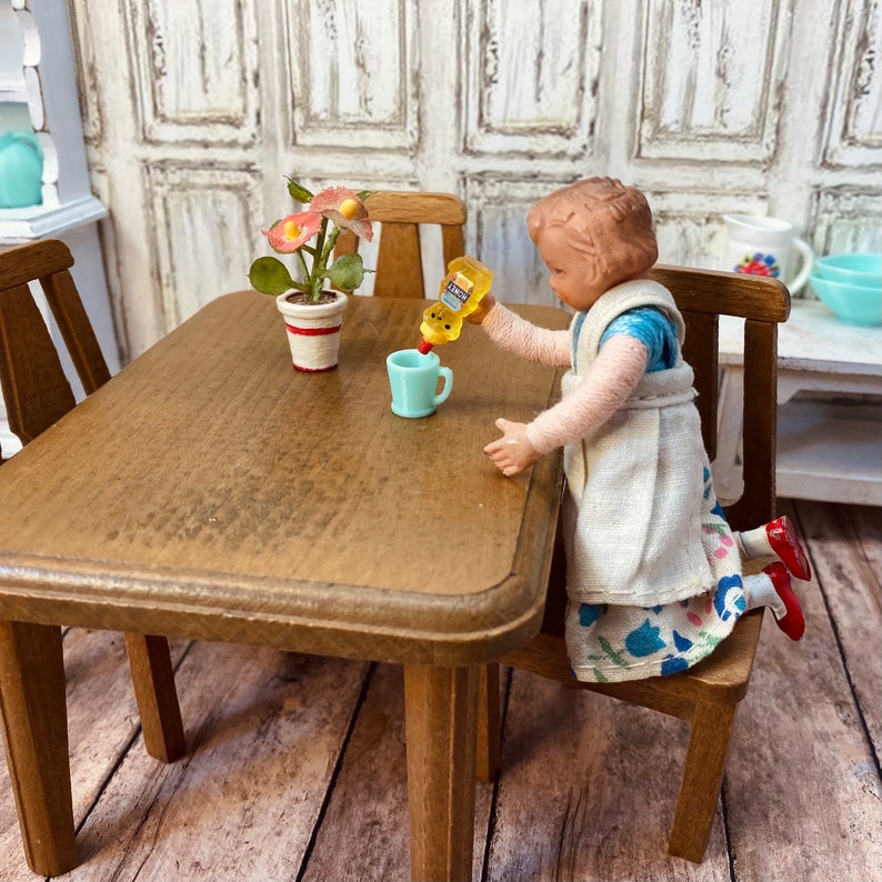 May include: A doll wearing a white dress with blue floral print is pouring yellow liquid from a bottle into a blue cup. The doll is sitting at a wooden table with a potted plant with pink flowers.