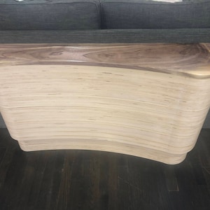 May include: A modern, curved wooden console table. The top is a dark, rich brown, while the body is a light, layered wood. The table has a unique, flowing design, perfect for an entryway or living room. The table is approximately 60 inches long.