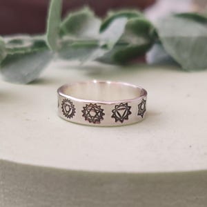 Può includere: Un anello d'argento con simboli dei chakra incisi. L'anello presenta una serie di disegni geometrici stilizzati attorno alla sua fascia. L'anello è esposto su una superficie verde chiaro, con un fogliame verde sfocato sullo sfondo.