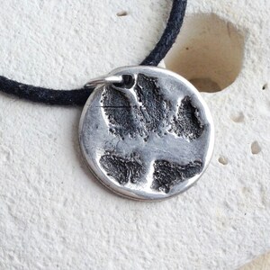 May include: A silver pendant necklace with a paw print design. The pendant is round and has a textured surface. The necklace is made of black cord.