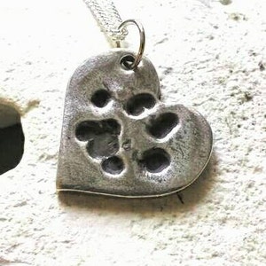 May include: A silver heart-shaped pendant necklace with a paw print design. The pendant is made of metal and has a textured surface.