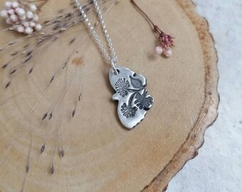 Op de afbeelding: Een zilveren ketting met een delicate hanger met een bloemenmotief. De hanger is gemaakt van metaal en heeft een getextureerd oppervlak. De ketting ligt op een houten oppervlak.