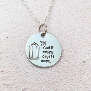 May include: Silver necklace with a round pendant featuring a bird flying out of a cage and the text "until every cage is empty".