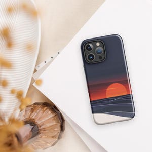 Puede incluir: Una funda de teléfono negra con una escena de puesta de sol y océano. La funda tiene un fondo azul oscuro con una puesta de sol roja y naranja y un océano azul. La funda del teléfono tiene un recorte para el objetivo de la cámara.