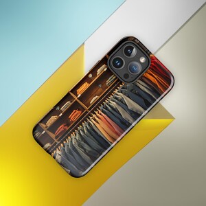 Op de afbeelding: Een telefoonhoesje met een foto van het interieur van een kledingwinkel. Het hoesje toont een close-up van een kledingrek met een selectie van kleurrijke shirts en jassen die aan hangers hangen. Het hoesje heeft een zwarte rand en een uitsparing voor de camera lens.